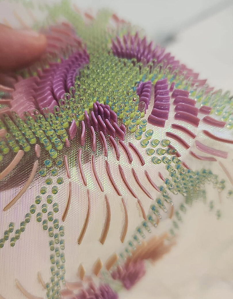 Close-up of intricate bead and sequin embroidery in purple, green, and pink hues on translucent fabric. A hand is partially visible holding the material.