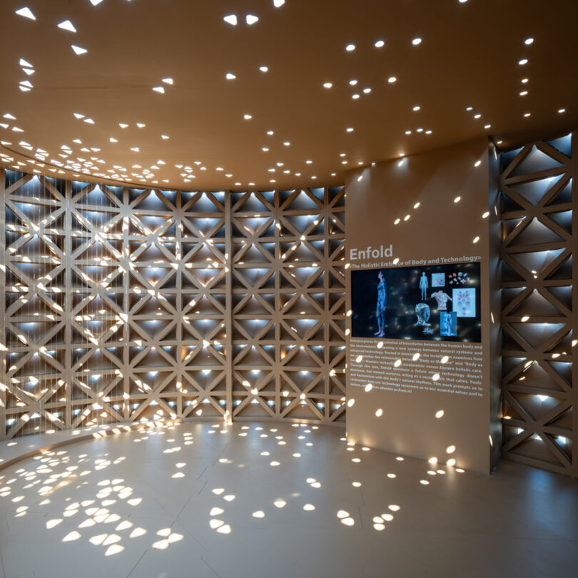 A room with geometric-patterned walls and ceiling, creating star-like light patterns. A screen and text titled "Enfold" are displayed on one wall.