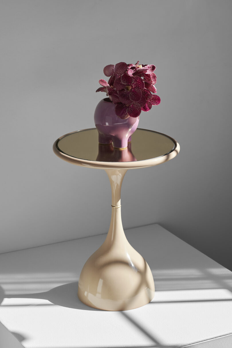 A modern beige vase from Entler Design, with dark pink flowers, graces a metallic round table featuring curvy support. This elegant planter is perfectly situated in a sunlit room, casting intriguing shadows.