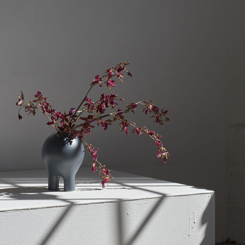 A gray Entler Design planter with four legs holds wilted red flowers, placed on a white surface with shadows cast in a dimly lit room.