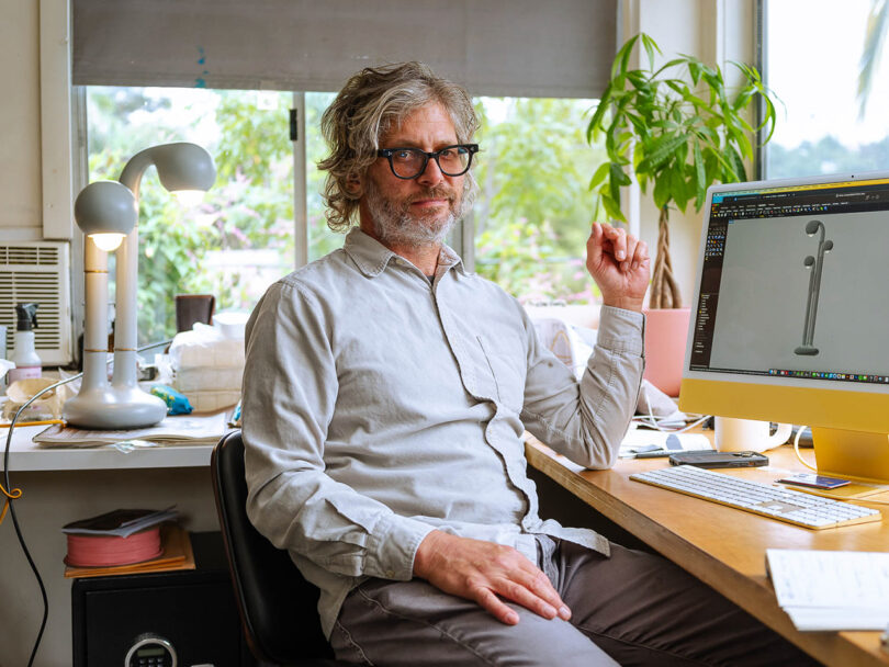 A person with glasses and a beard sits at a desk with a computer displaying an Entler Design 3D model. A planter and various desk items are in the background, adding charm to the creative workspace.