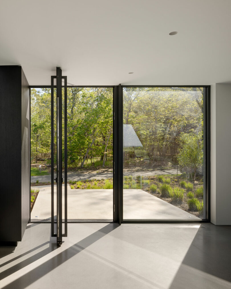 Modern interior with an open glass door leading to a patio, showcasing a view of a landscaped garden with green trees.