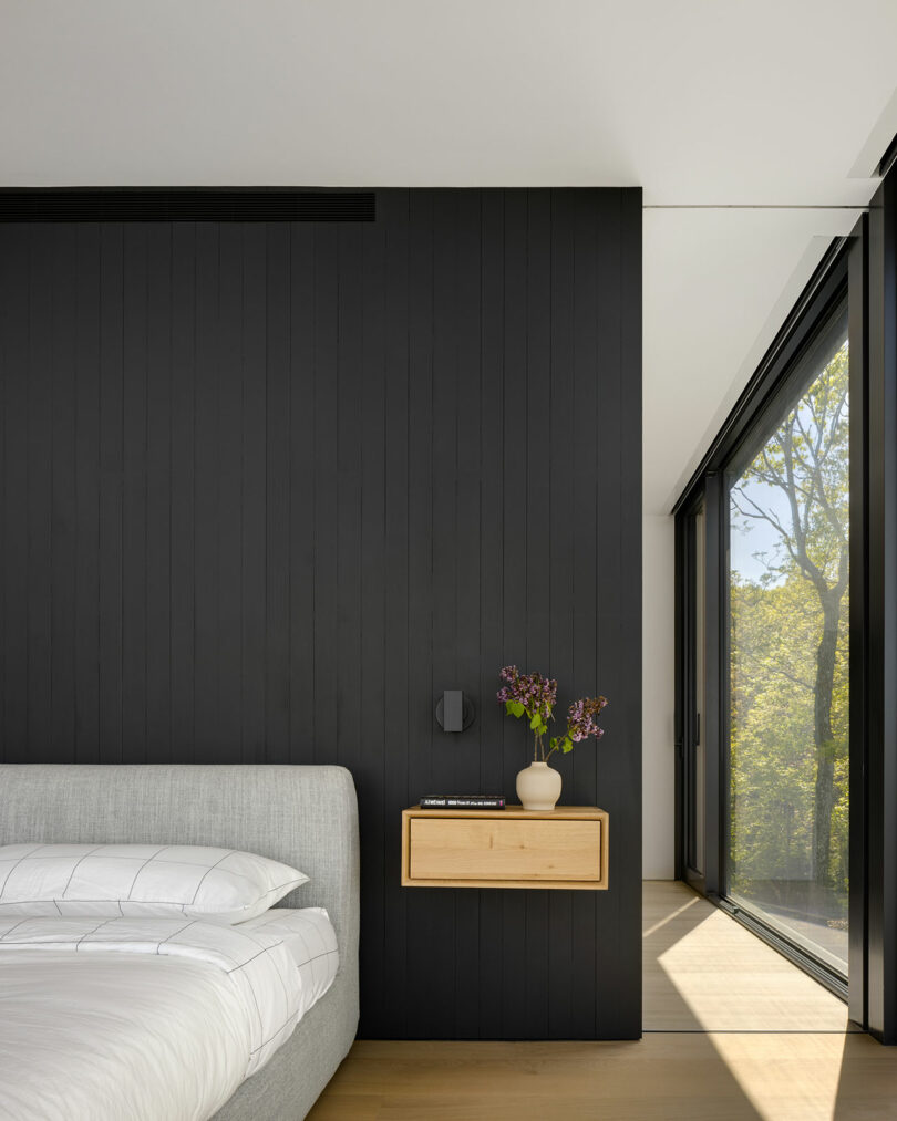 Bedroom interior with black accent wall, gray bed, and wooden floating nightstand. A vase with flowers is on the nightstand. Large window with a view of trees to the right.