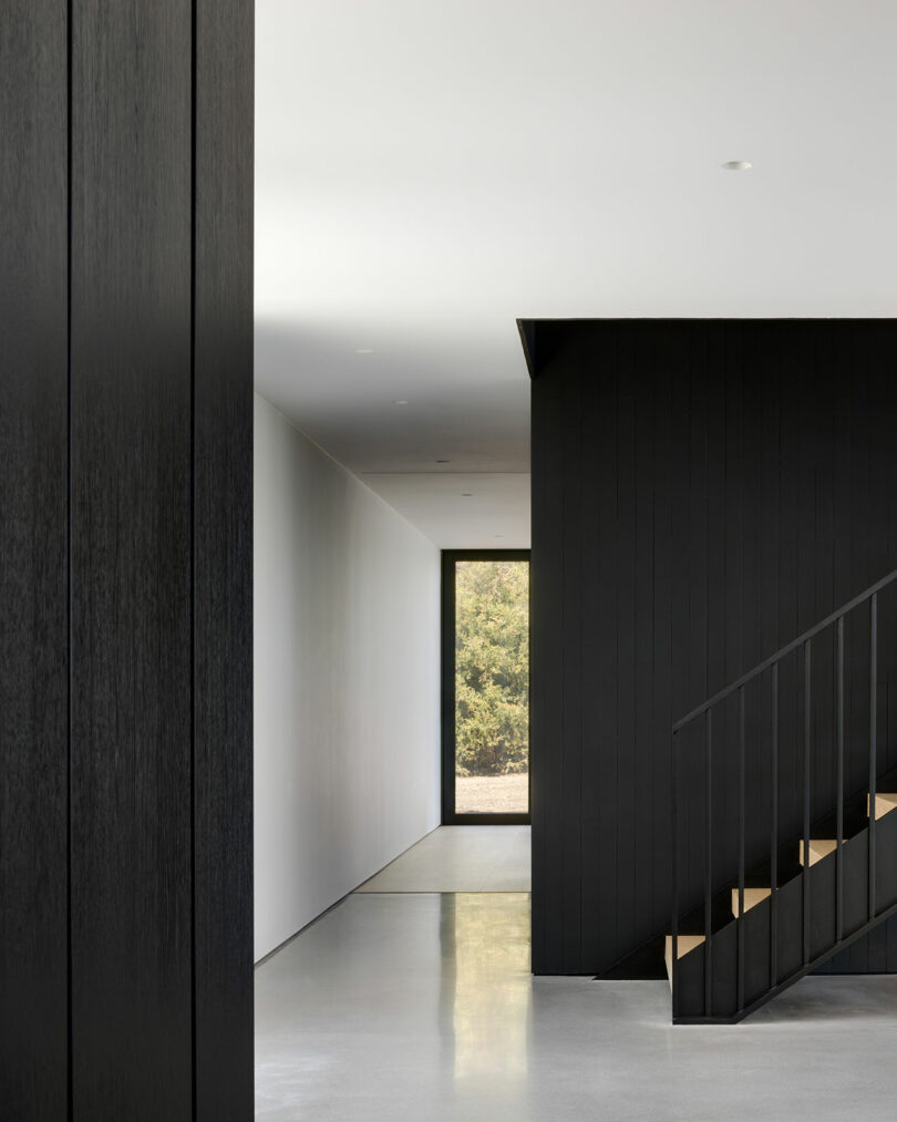 Minimalist modern interior with black vertical paneling and staircase. A hallway leads to a large window with a view of greenery. The floor is polished concrete.