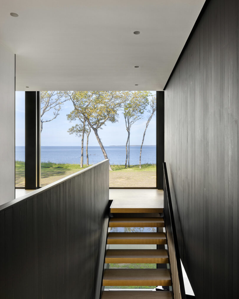 Interior hallway with wooden stairs leads to a large window, revealing a view of trees and a calm sea under a clear sky.