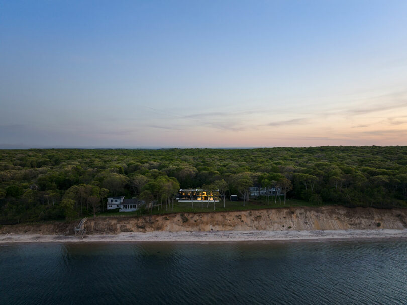 Aerial view of a coastal landscape at sunset with forested area, a few houses near the edge of a sandy cliff, and a calm body of water in the foreground.