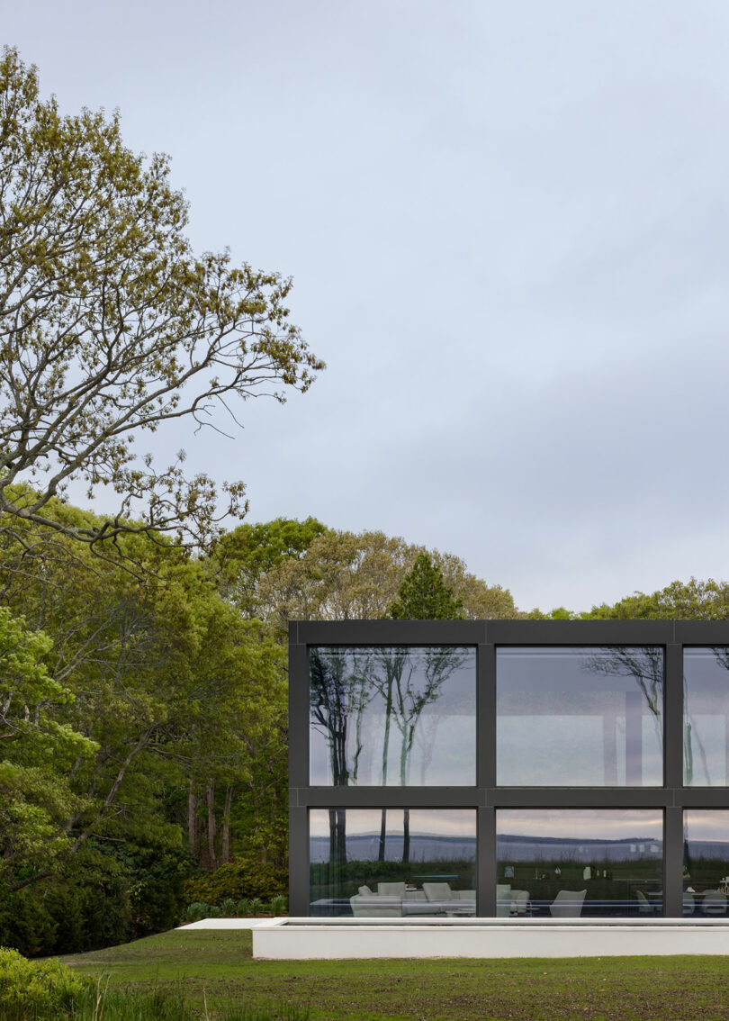 Modern glass house surrounded by trees with reflections in the windows, set against a cloudy sky.