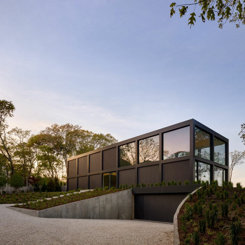 Modern black rectangular house with large windows, surrounded by trees and a gravel driveway, under a clear sky.