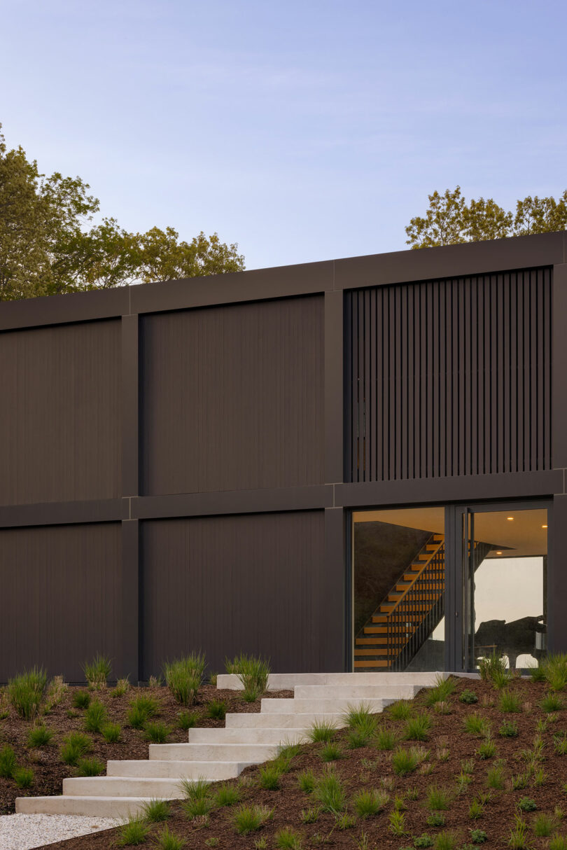 Modern building with dark vertical panels and large glass doorway, set against a clear sky. Steps lead up through landscaped greenery.