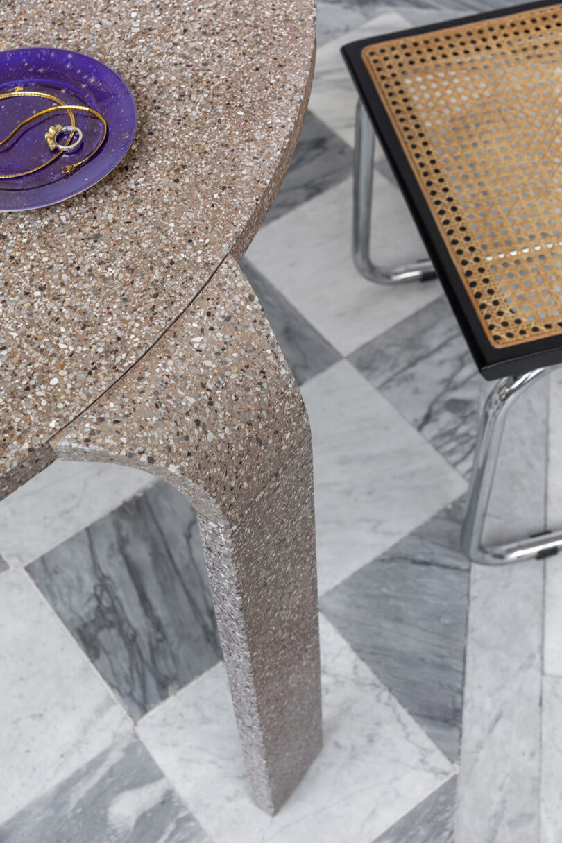 A terrazzo table with a purple dish and gold jewelry sits on a geometric marble floor next to a cane-backed chair.