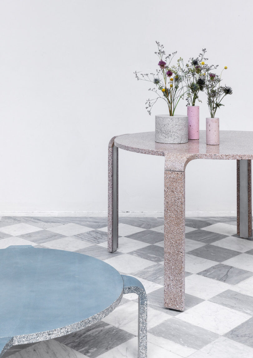 Two terrazzo tables on a checkered floor, one blue and round, the other pink and taller with curved legs, topped with vases containing assorted flowers.