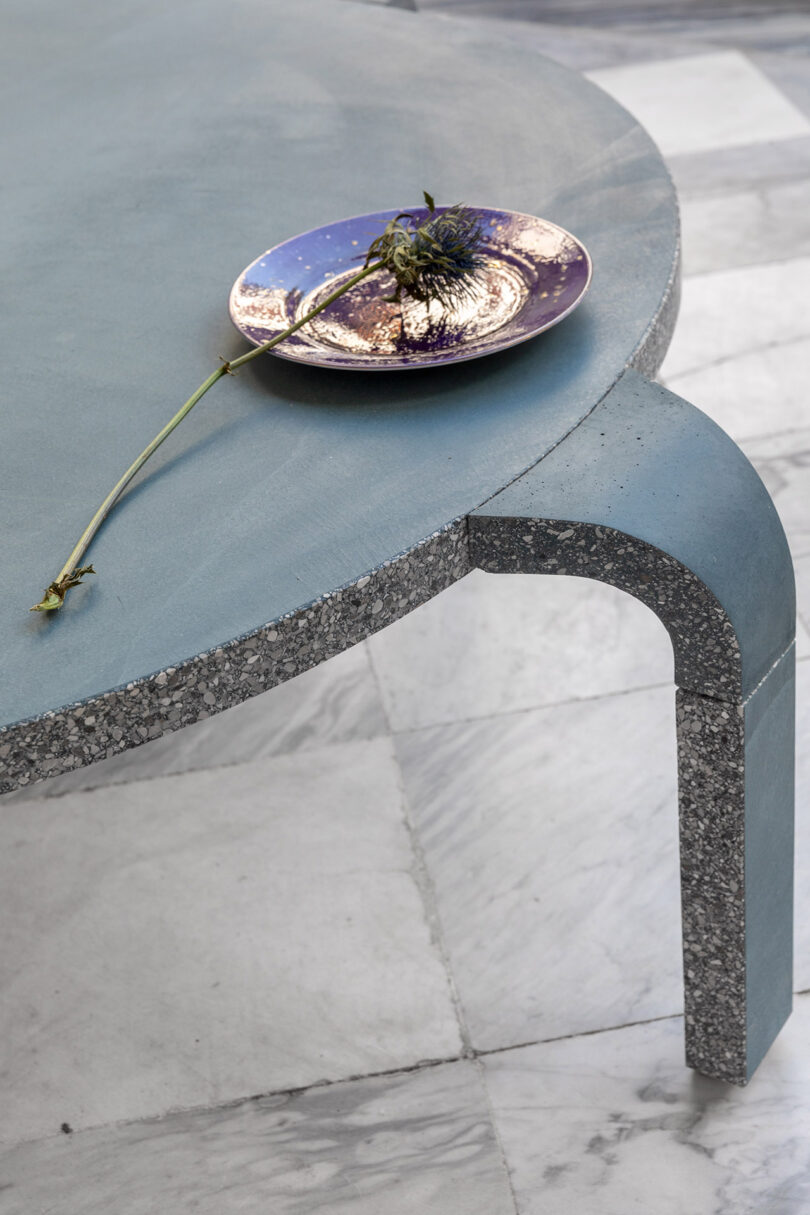 A round table with a terrazzo edge holds a small, decorative plate with a single wilted flower on a marble floor.