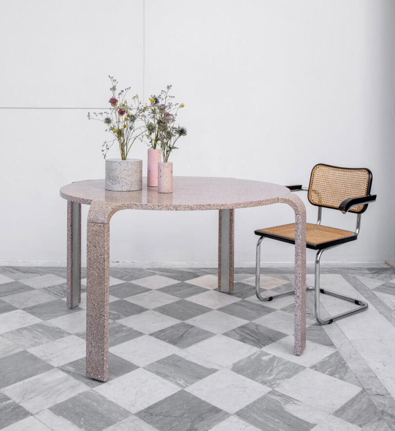 A round terrazzo table with modern legs holds two vases of flowers and two small cylindrical containers. A cane and metal chair is positioned beside the table on a checkered floor.