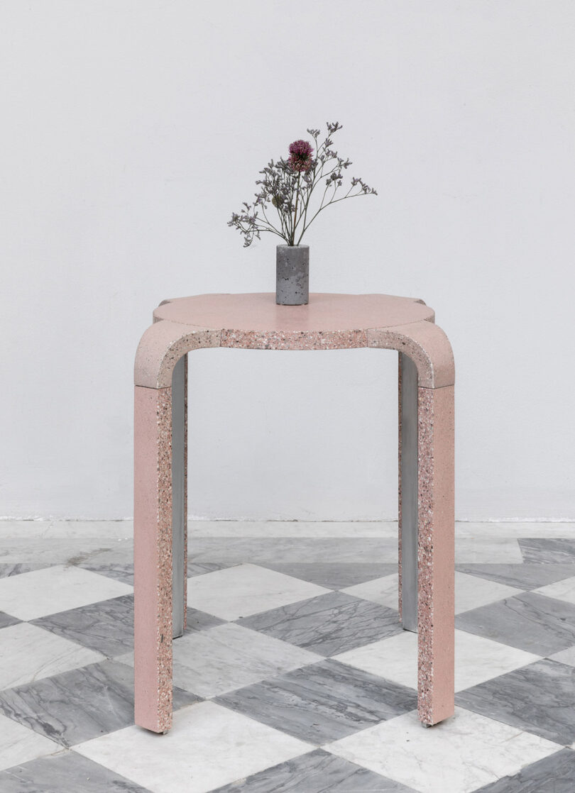 A small table with a pink concrete design holds a vase with dried flowers against a white wall. The floor features a checkered black and white pattern.