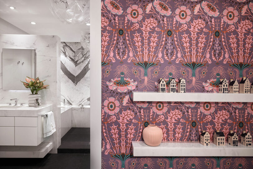 Bathroom with white cabinetry and floral patterned wallpaper, featuring shelves with miniature house models and a pink vase.