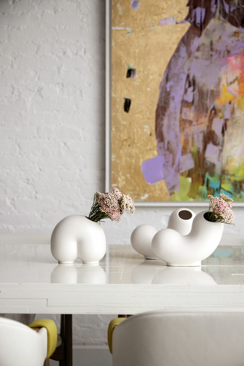 White abstract vases with flowers on a glossy table, set against a white brick wall and colorful abstract artwork.