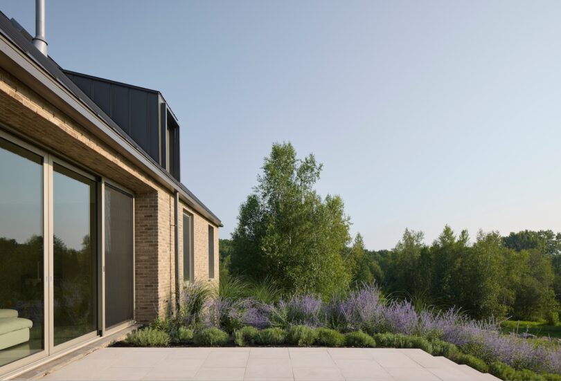 Modern brick house with large windows overlooking a garden with purple flowers and trees under a clear sky.