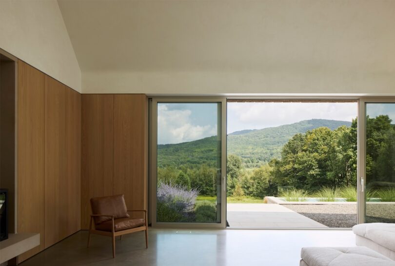 Modern living room with a leather chair, large sliding glass doors, and a view of green hills and trees. A gray floor and light wood wall complete the minimalist design.