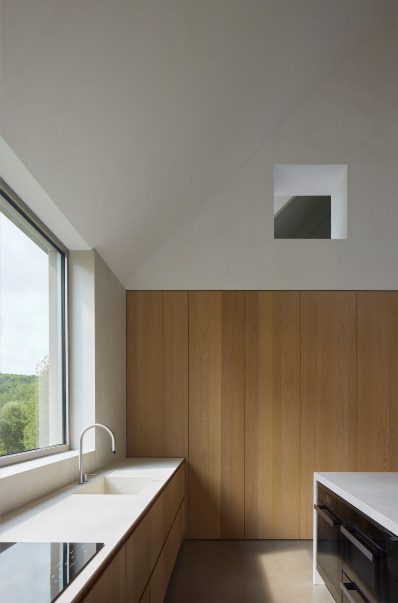 Minimalist kitchen with light wood cabinets, white countertops, an induction cooktop, and a large window offering a view of greenery. A small square window is near the ceiling.