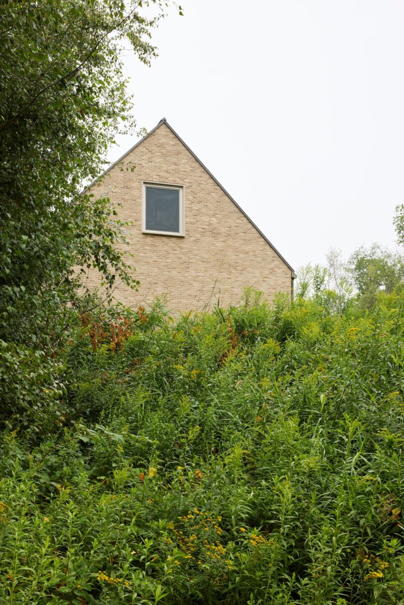 A brick house with a single window on the upper level, partially obscured by lush greenery and trees.