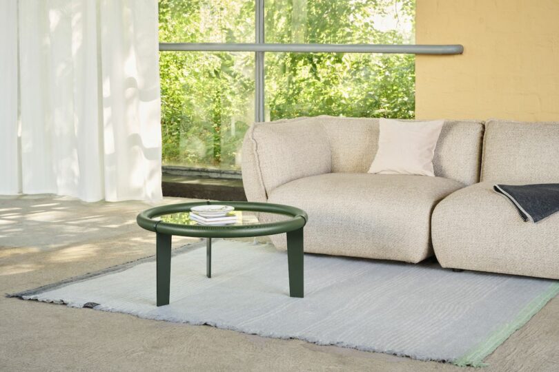 A beige sofa with cushions and a dark green coffee table on a light gray rug in a sunlit room with large windows and a yellow wall