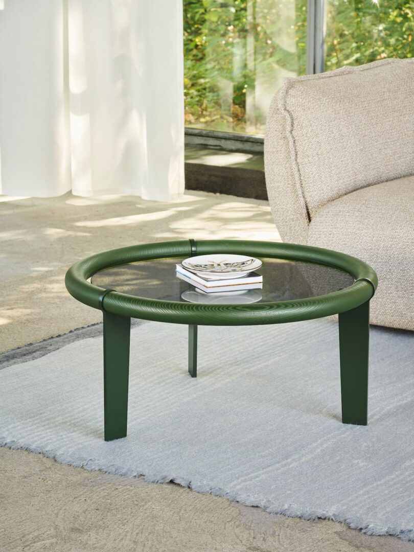 A round glass-top coffee table with a green frame is on a light gray rug next to a beige sofa. A small stack of coasters sits on the table