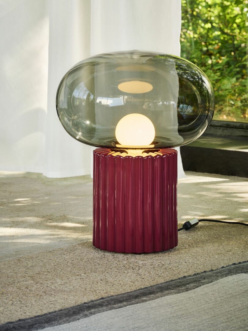 A modern table lamp with a red textured base and a large clear glass shade, placed on a light-colored surface near a window with greenery in the background