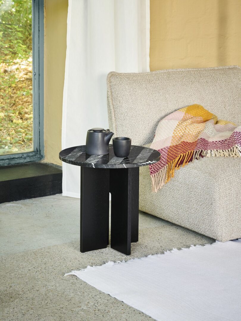 A small black table with a marble top holds a black teapot and cup next to a sofa with a colorful throw blanket. Nearby is a window with a view of greenery outside