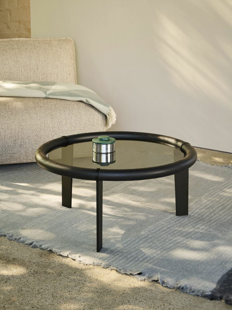 A modern black round coffee table with a glass top on a blue rug. A small metal container sits on the table, next to a beige sofa with a folded blanket draped over the arm