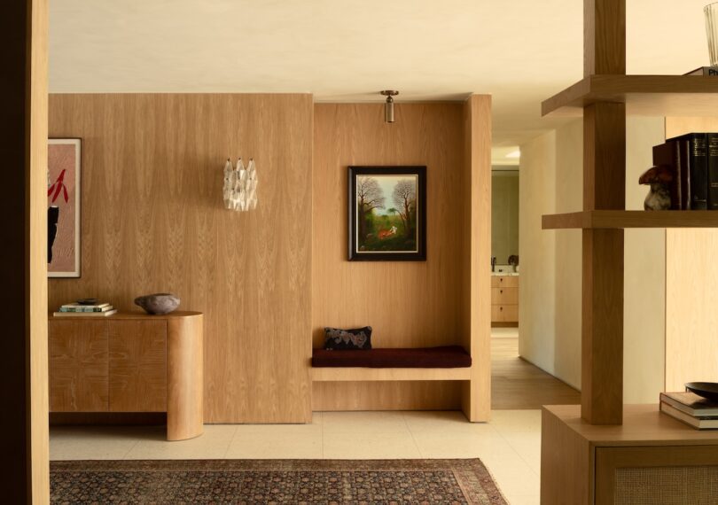 A modern interior with wooden walls, a bench with a cushion, a painting, and a side table with books. Shelving on the right and a rug on the floor complete the room.