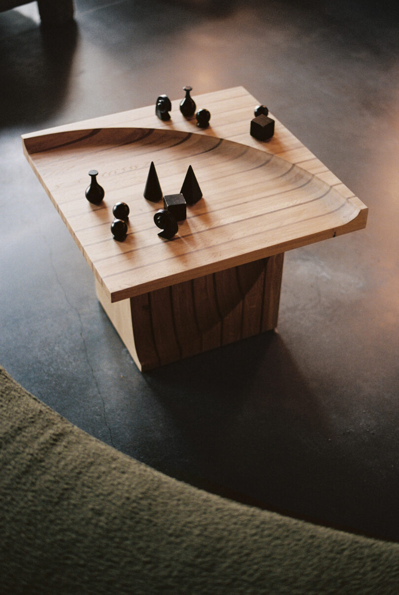 A wooden coffee table with various black geometric shapes is positioned on a dark floor. A green cushioned chair is partially visible in the foreground