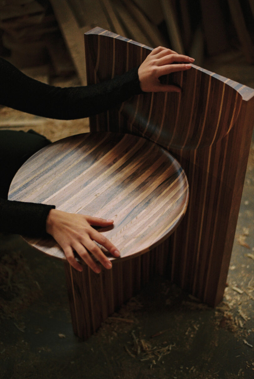 A person assembles a striped wooden chair with circular seat and angular backrest, surrounded by wood shavings