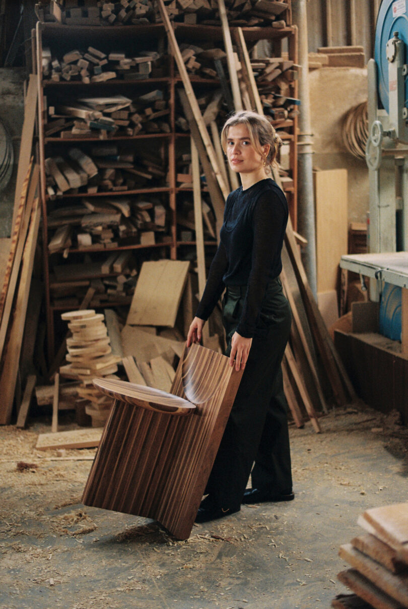 A person in a workshop holding a wooden chair piece. Shelves with stacked wood and tools are in the background