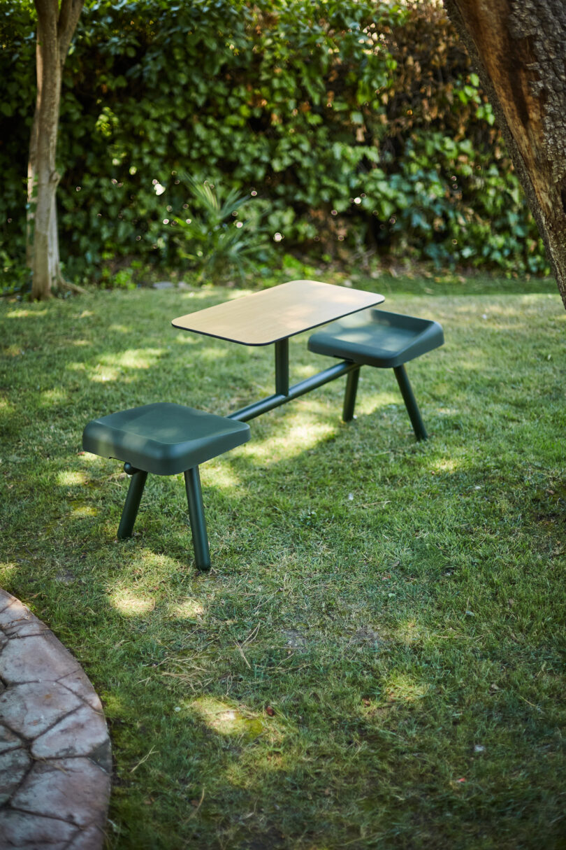 A small outdoor table with two attached green cushioned seats on a grassy area, surrounded by trees and shrubbery