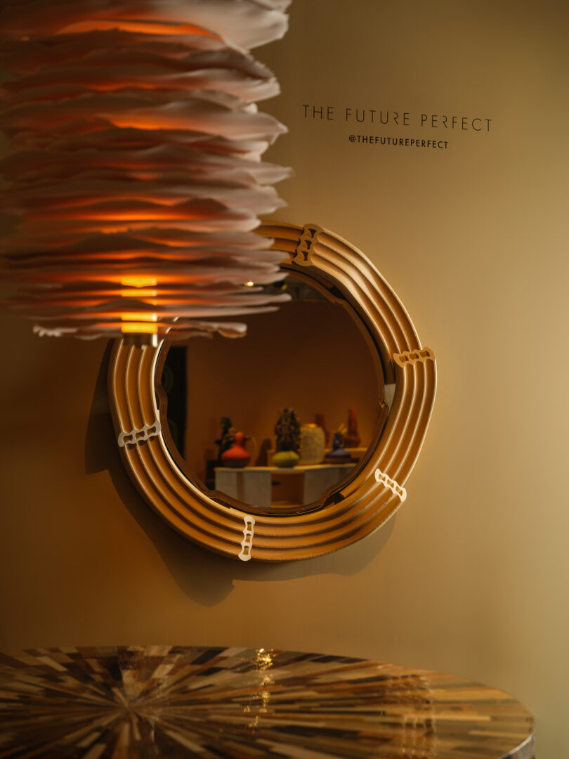 A round mirror with a unique frame is mounted on a wall. A layered light fixture hangs in the foreground. A blurred view of colorful decor items is reflected in the mirror