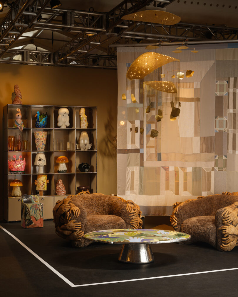 A room with two textured chairs, a patterned table, art pieces on a shelf, and a beige curtain with hanging lights