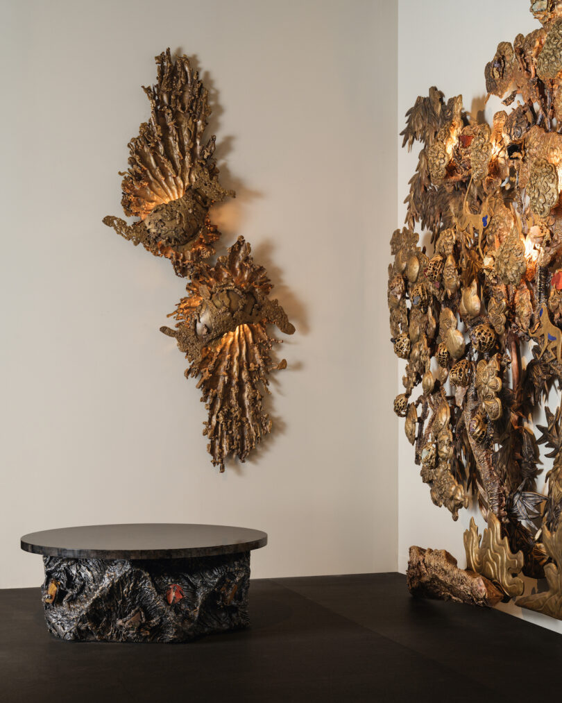 An ornate wall installation and a black sculptural table with intricate textures are displayed in a gallery setting