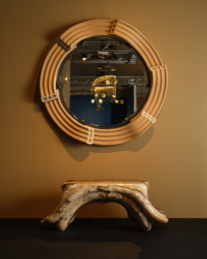 Round mirror with a wooden frame on a tan wall, reflecting a chandelier. Below is an abstract, marbled bench with curved legs