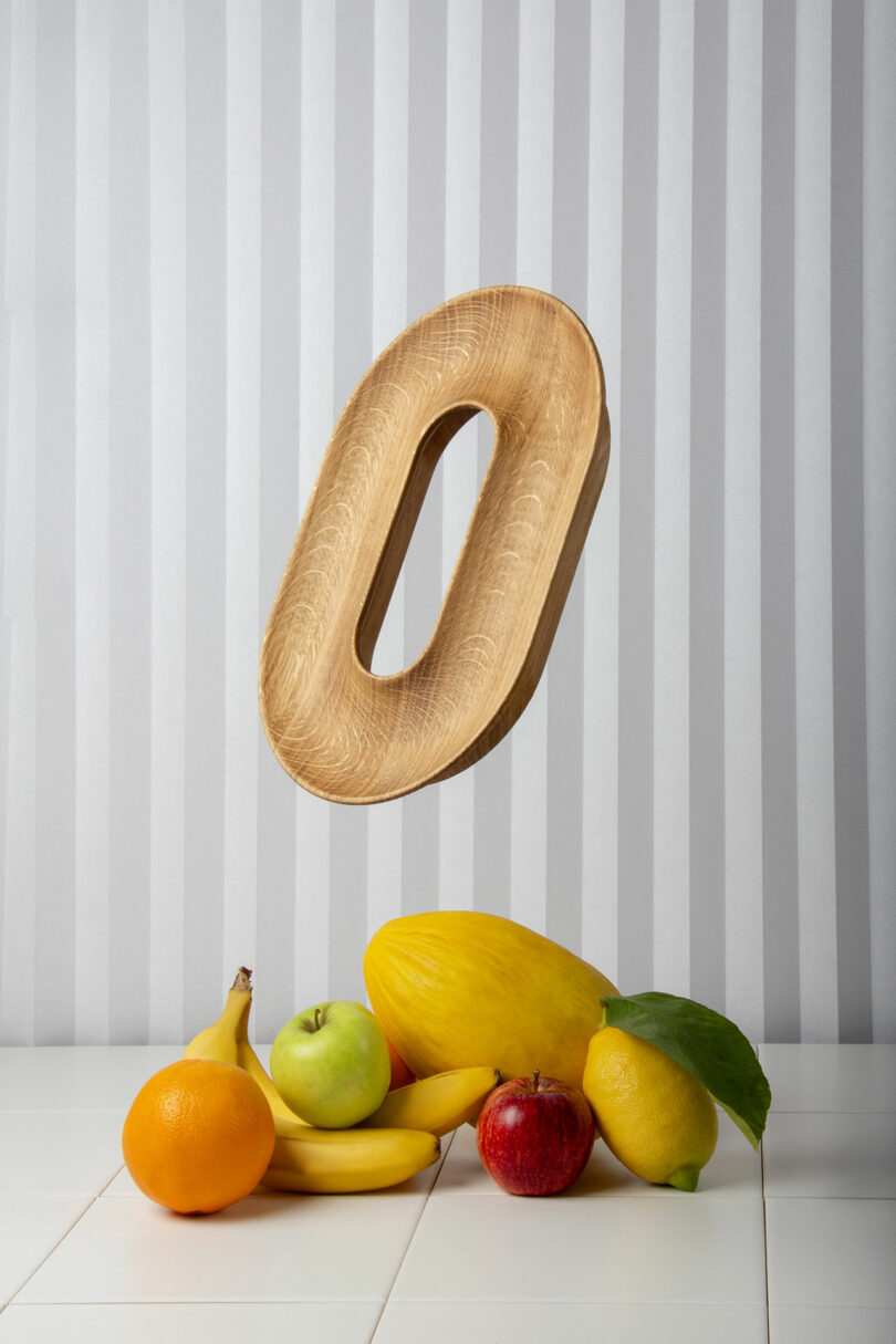 A wooden fruit vessel floats above a table with a variety of fruits, including bananas, an apple, an orange, a lemon, and a mango, against a striped backdrop