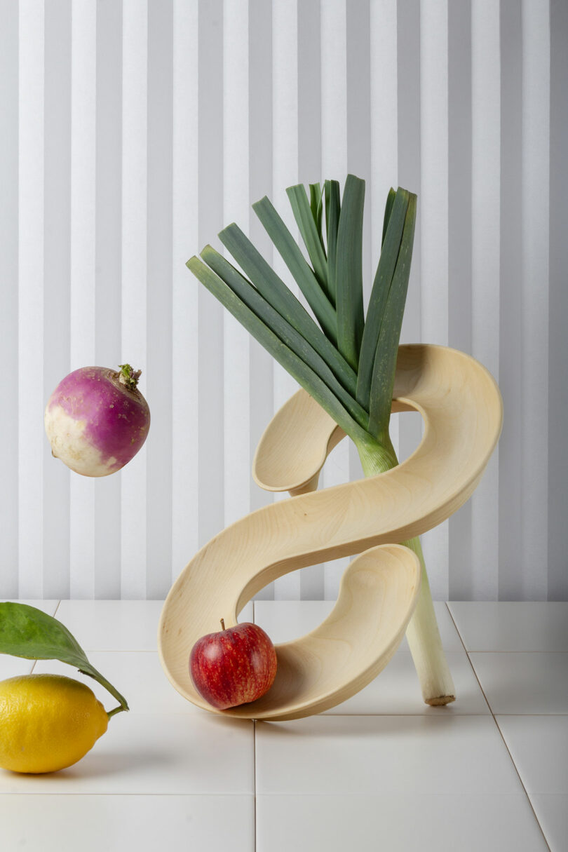 A wooden spiral stand holds a leek. An apple rests at the bottom, while a turnip appears to float mid-air. A lemon with attached leaves is nearby, set against a vertically striped background