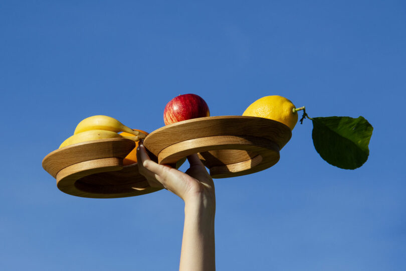 A hand holds wooden trays against a blue sky, displaying apples, bananas, and a lemon with a leaf