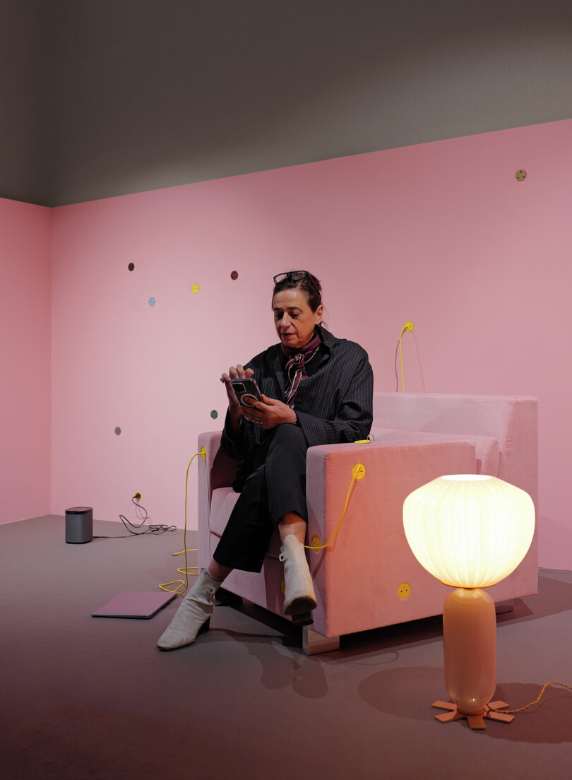 Person sitting on a pink chair in a room with pink walls, holding a phone. There's a lamp and various cables around.