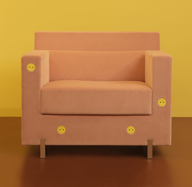 A beige armchair with smiley face buttons is set against a yellow background. The chair has a boxy design and metal legs.