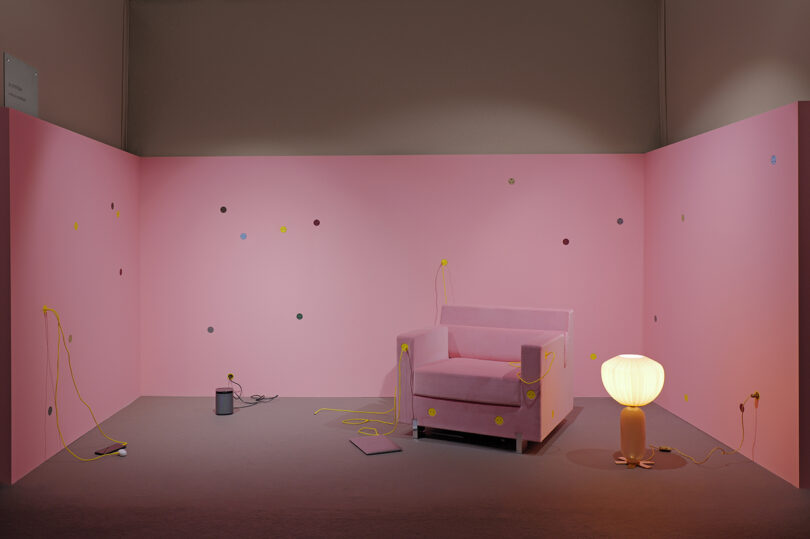A pink room with a pink armchair, a small pink side table, a paint can, a glowing lamp, and yellow cords. Multicolored dots are scattered on the walls.