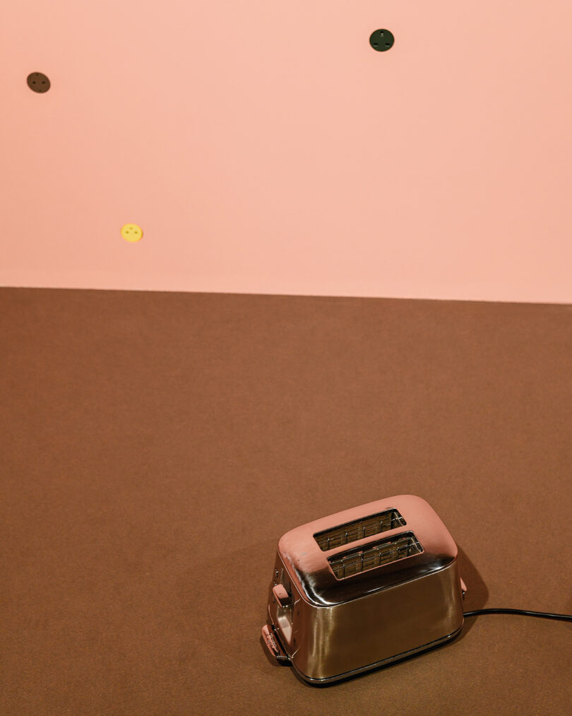 A silver toaster sits on a brown surface with a light pink wall in the background, featuring a few scattered, colored round stickers.