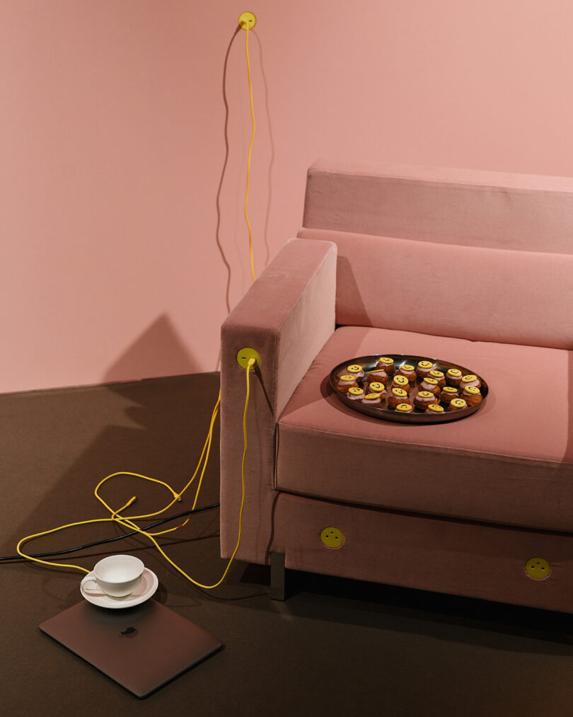 A pink sofa with yellow plugs and cords, holding a tray of small desserts, is placed on a brown floor. A laptop and a cup on a saucer sit nearby.