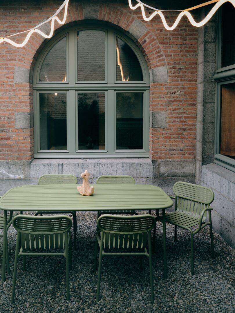 Outdoor dining set with six green chairs and a rectangular table, featuring a small animal sculpture, situated on gravel in front of a red brick wall and arched window. String lights are above
