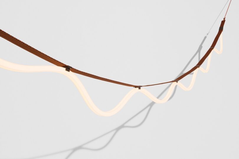 Curved light fixture with a wavy design suspended by a brown strap against a white background. Shadows cast on the wall