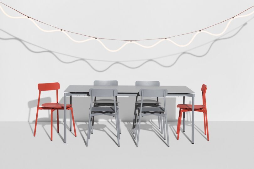 A minimalist dining set with a black table, six chairs—four gray, two red—and overhead wavy strip lights against a plain white background