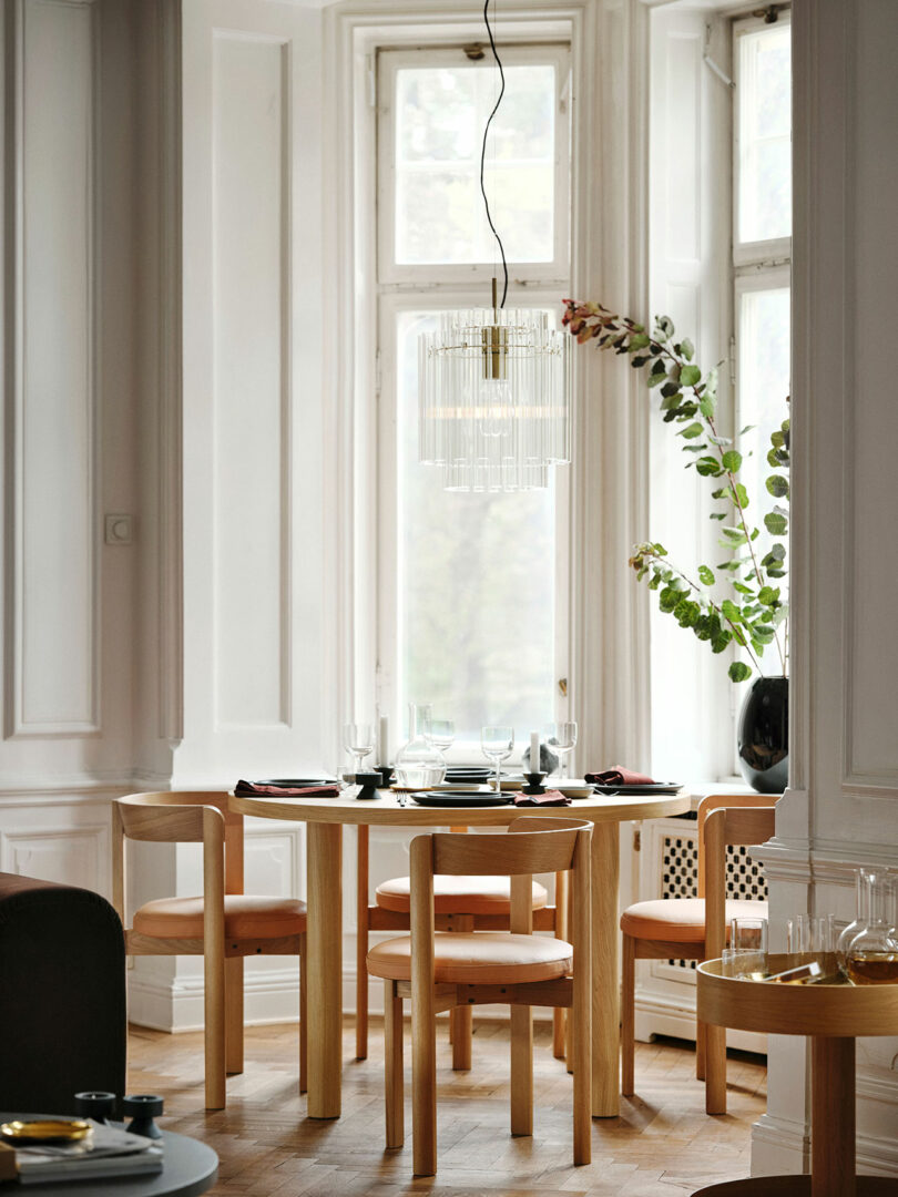 A small round Stockholm IKEA dining table with four wooden chairs is set near tall windows, with a modern chandelier above and a vase with greenery on the windowsill.